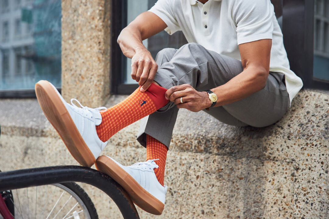 A man sits on a concrete ledge, wearing grey trousers, a white polo shirt and white sneakers. He reaches forward to pull up his russet orange socks by London Sock Company.