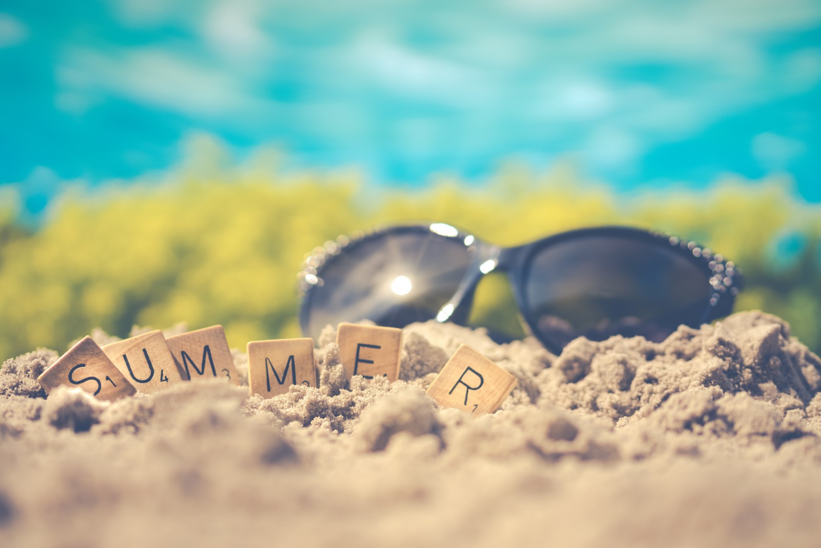 A pair of sunglasses in the sand on a beach, with scrabble letters spelling out summer. 