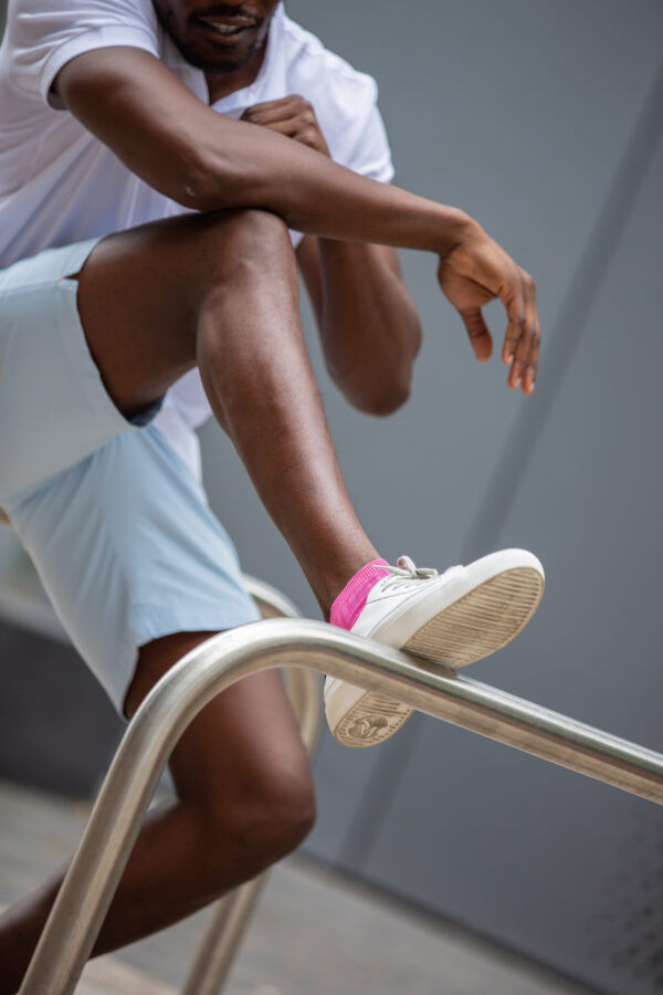 A black man wearing a white polo shirt, blue shorts, white trainers and pink trainer socks from London Sock Company rests his foot on a metal bar