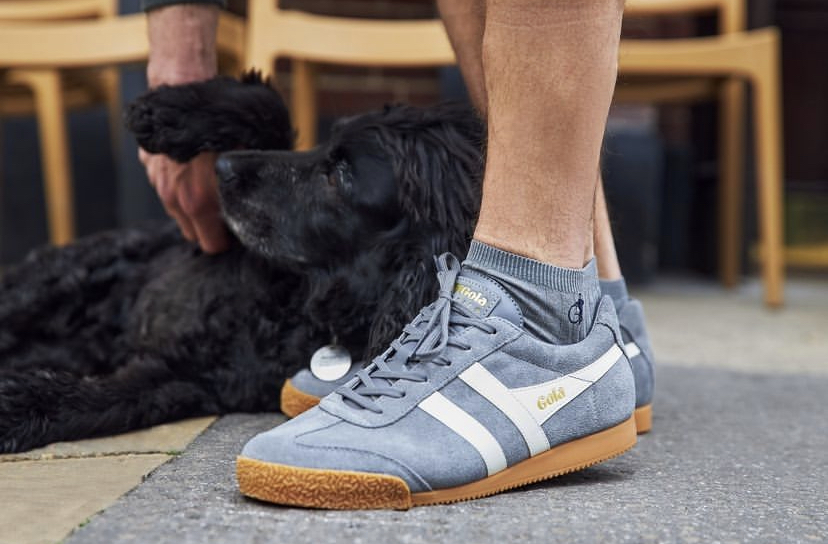 A close up of a man's foot in grey socks and grey trainers. His hand is in shot, stroking a black Spaniel dog.