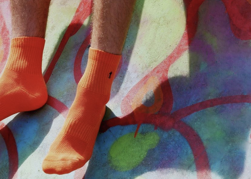 A man's feet in orange cotton sports socks from London Sock Company hang down on a wall that is covered in neon graffiti.