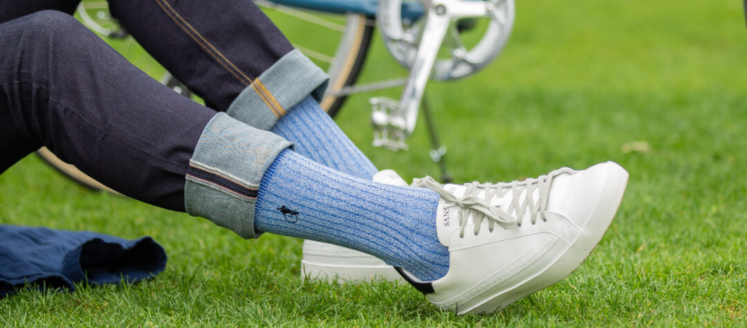 A man in jeans stretches his leg out in front of his parked bike, wearing blue marl socks from London Sock Company and white trainers