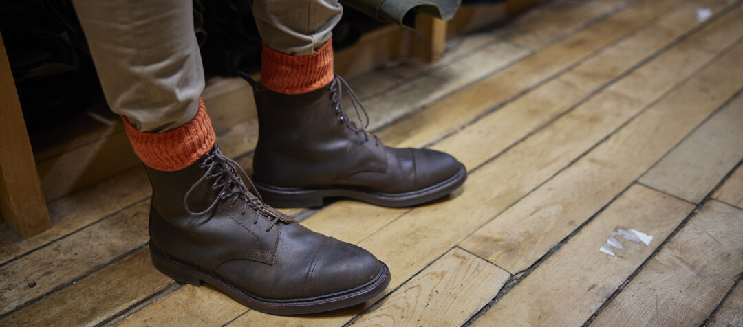 A man's feet on a wooden floor, wearing brown leather lace-up boots from Crockett & jones and spiced orange socks from London Sock Company.