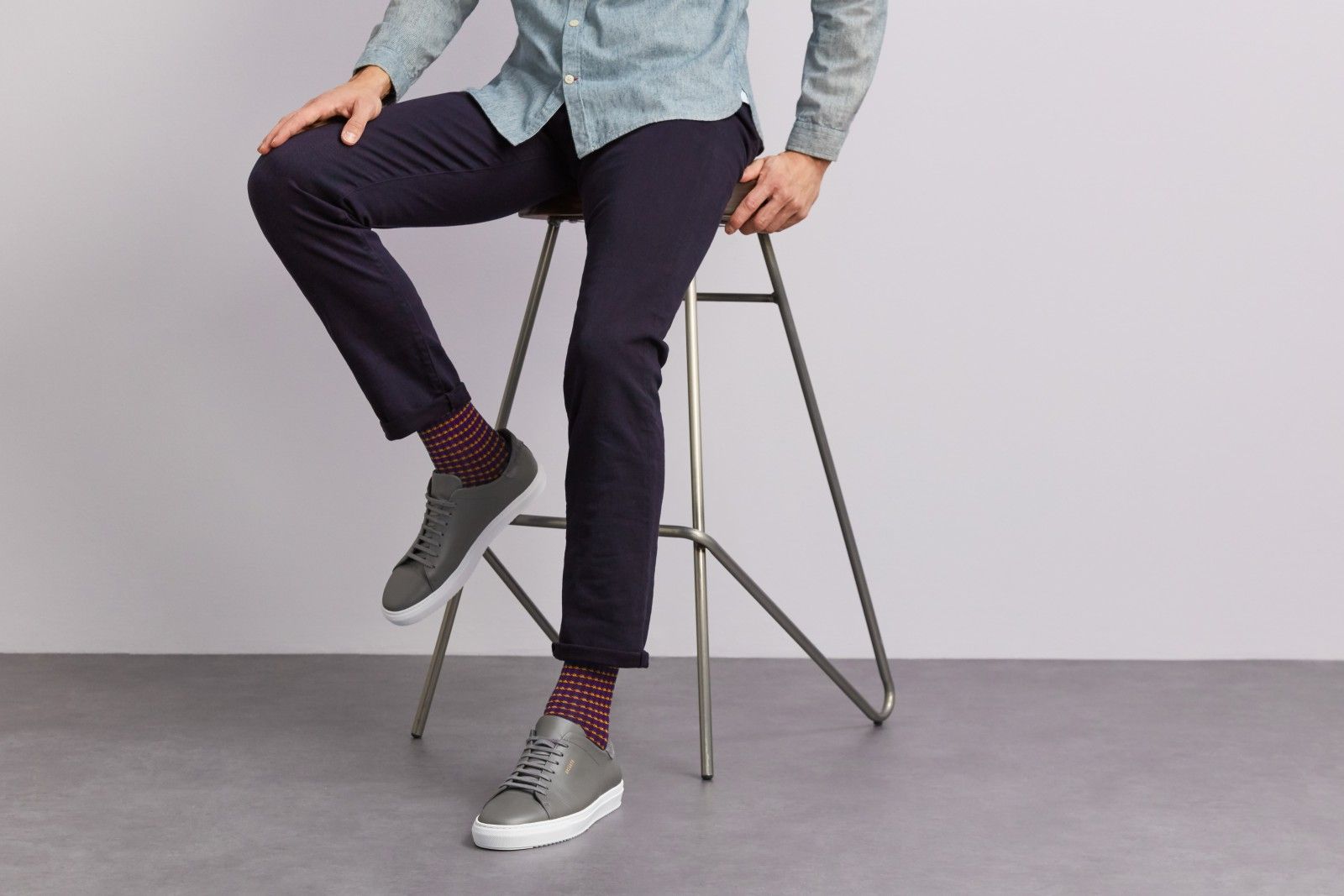 A man sitting on a stool wearing a light blue shirt, navy trousers, red pattern socks from London Sock Company, and grey trainers.