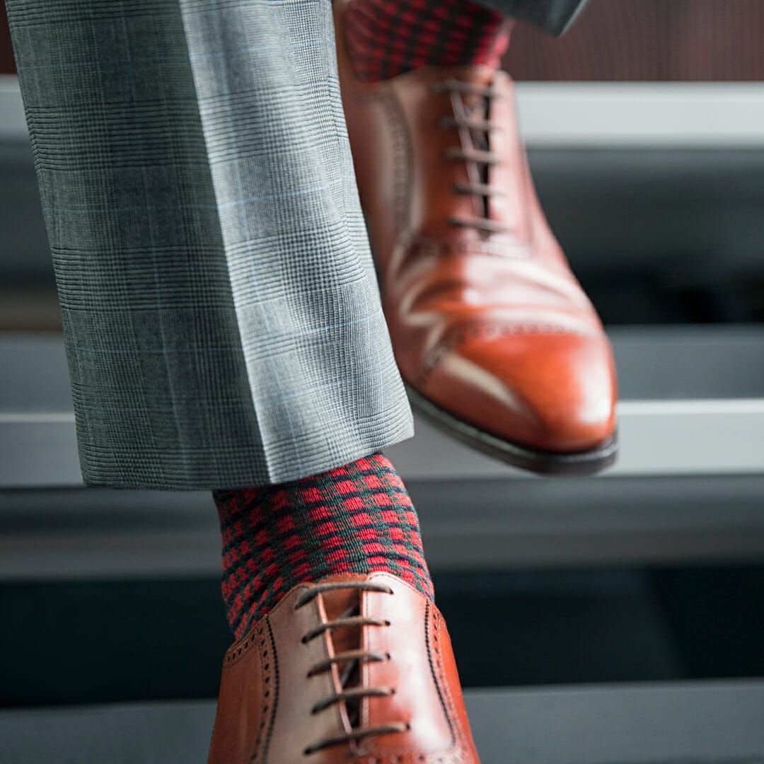 A man wearing a red sock with Houndstooth pattern, styled with a grey trouser and brown brogue.