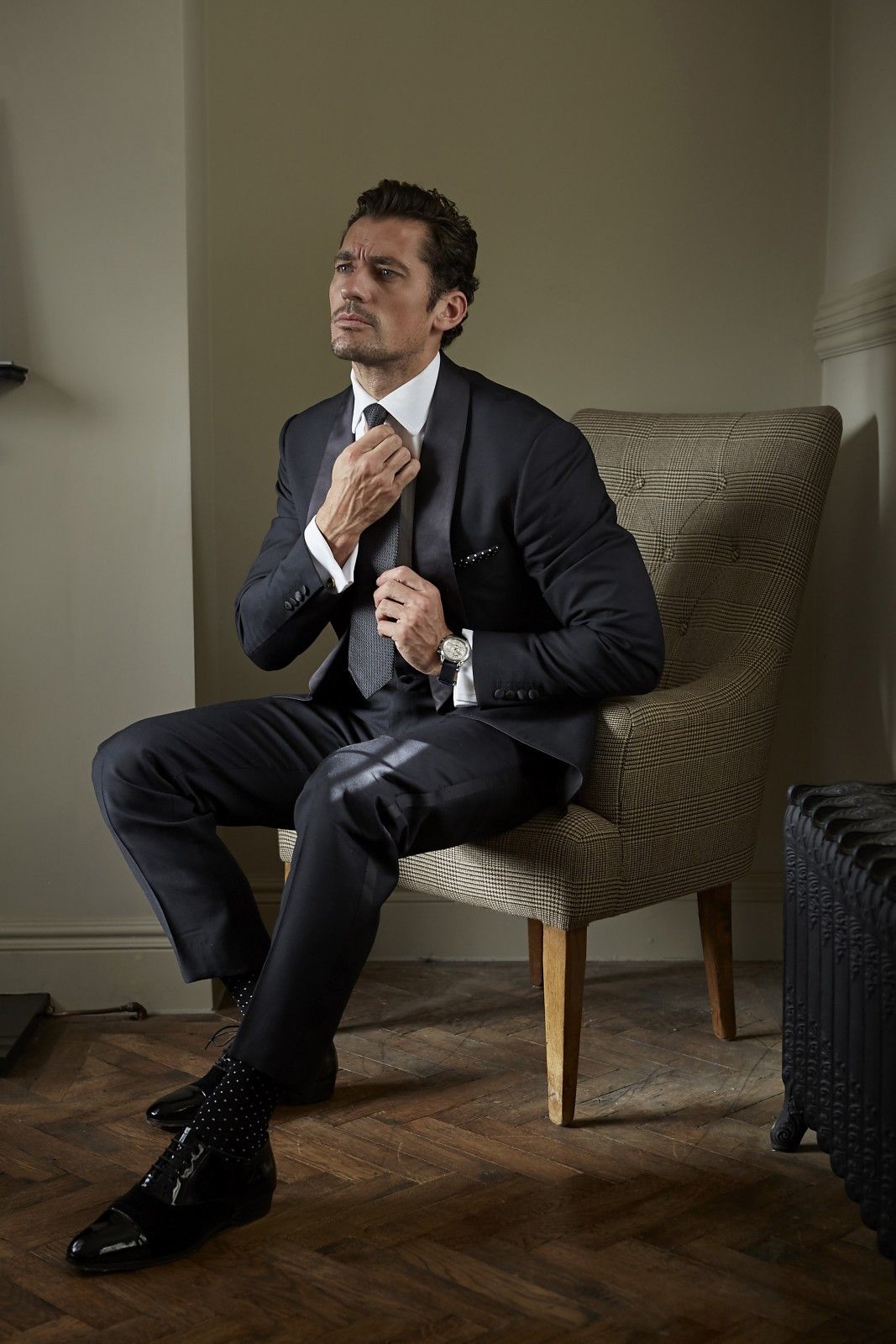 Model David Gandy tightens his tie while sitting on a leather chaior, wearing black tie and socks by London Sock Company.
