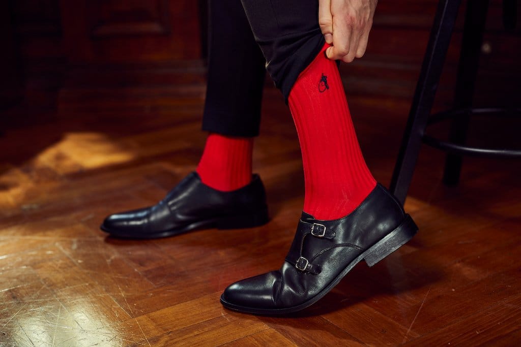 Photo of a man’s lower legs, wearing smart black trousers with bright red
socks and black leather monk strap shoes, pulling up the left sock with his left hand.