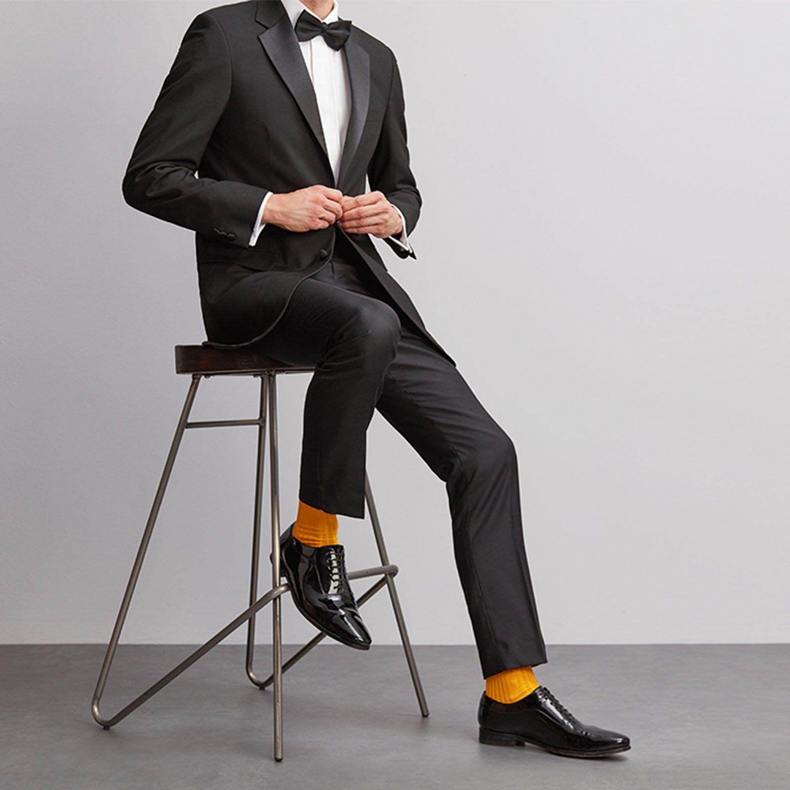 Man sitting on a stool wearing a tuxedo, bow tie and saffron yellow socks from London Sock Company.