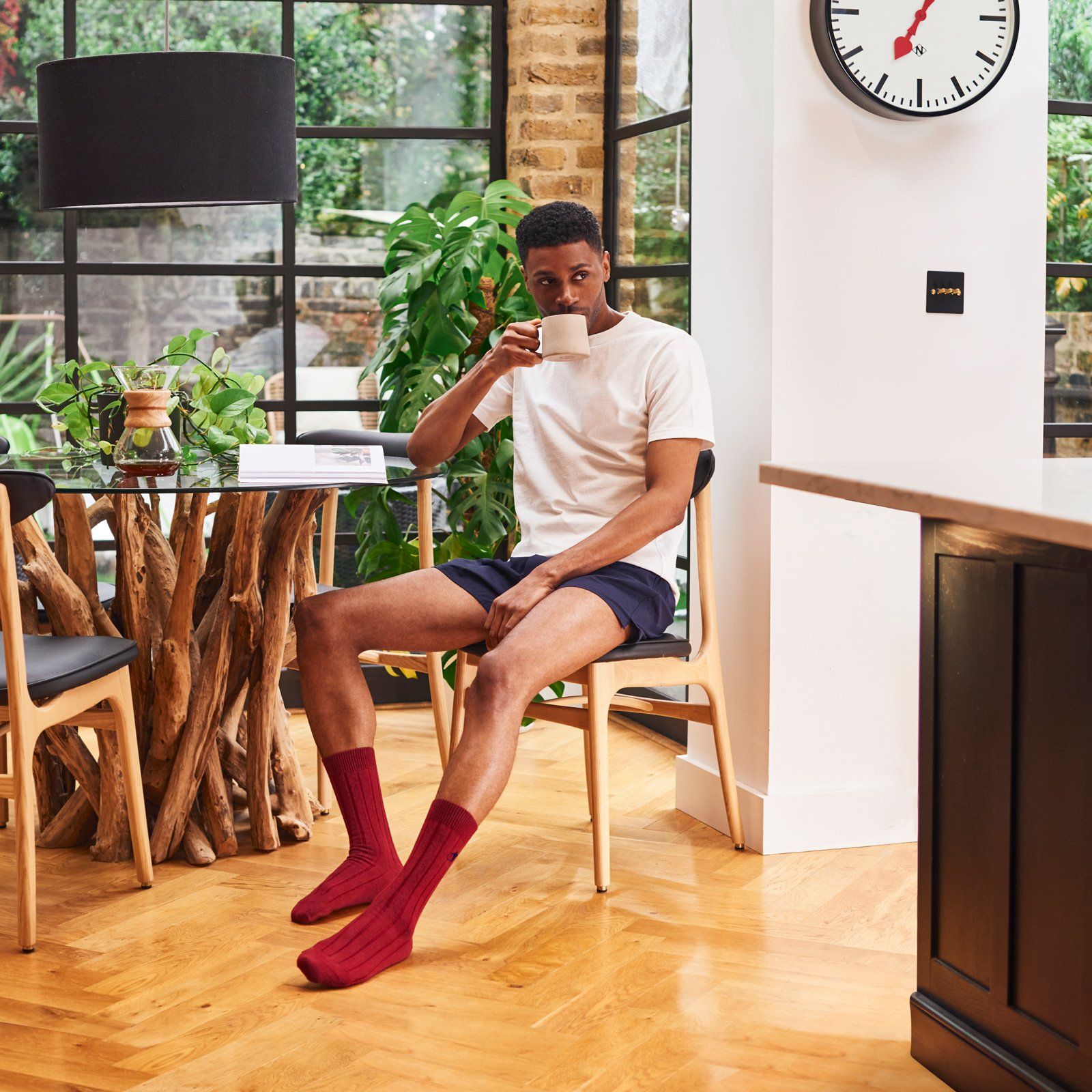 A man sitting on a chair in a kitchen holding a cup of coffee, wearing red
cashmere socks from the London Sock Company.