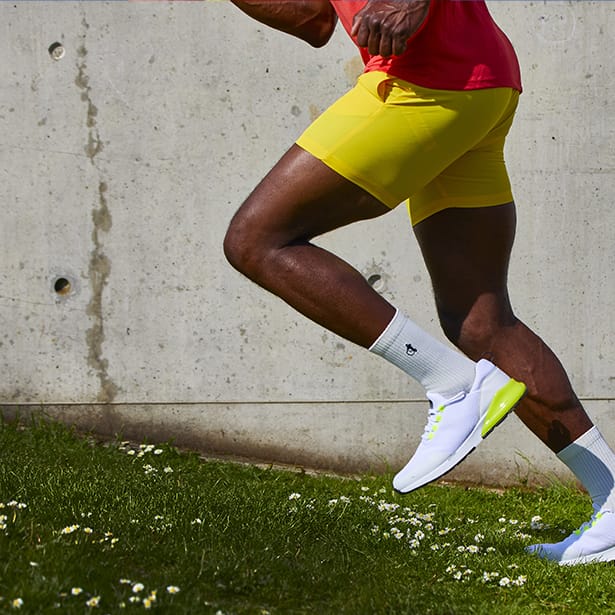 A black man wearing bright yellow shorts and a red tshirt with white sports socks by London Sock Company and white trainers runs up a grassy hill. Everything is very vibrant.