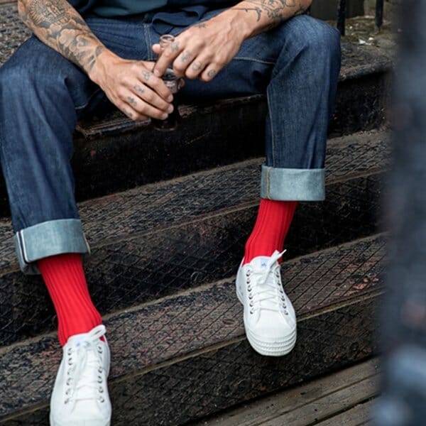 A picture of the bottom half of a man with tattooed arms sitting on some steps, wearing dark jeans with turned up cuffs, bright red socks from London Sock Company and white trainers.