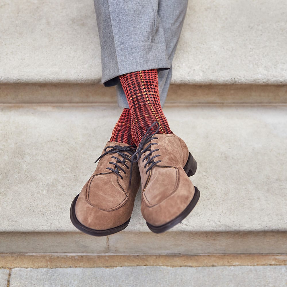 Close up on a man's feet, crossed over against a stone wall, wearing brown suede shoes, grey trousers and a pair of checked burnt orange socks by London Sock Company.