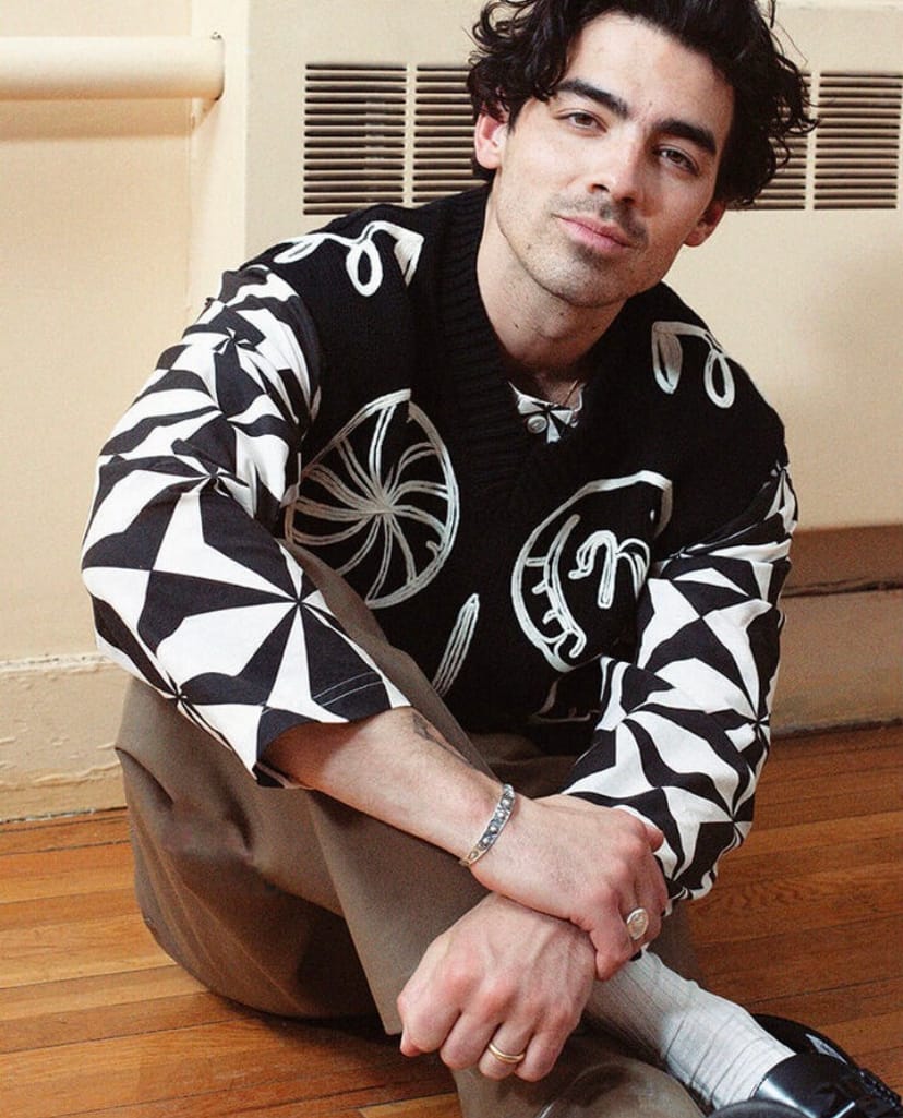 Singer Joe Jonas sits on the floor in a black and white patterned sweater and stone trousers, with his legs crossed. He wears white socks from London Sock Company and black loafers.