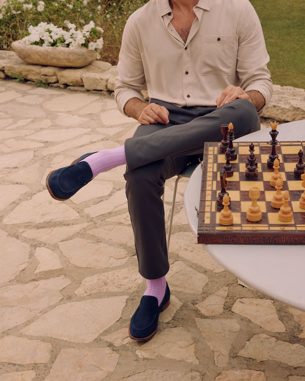 A man sits on a chair on a patio cross legged next to a chess board wearing grey chinos, blue suede loafers and lavender purple socks by London Sock Company.
