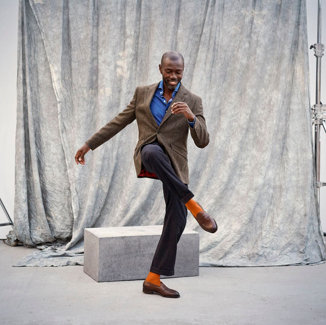 A man wearing smart tailored wool trousers, a brown tweed jacket and blue collared shirt dances, showing off his rust orange socks by London Sock Company.