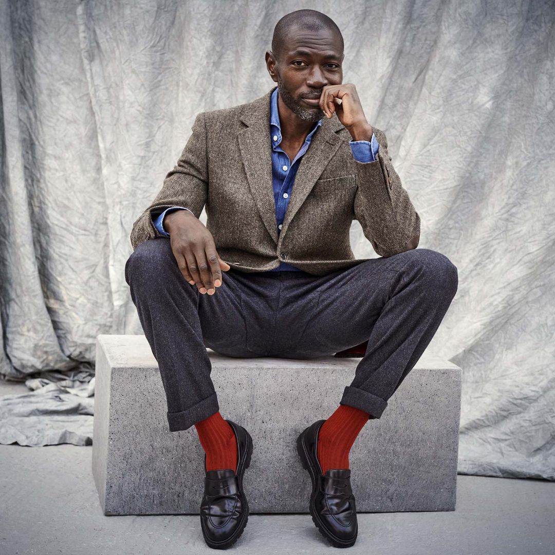 Photo of a man sitting on a grey concrete block wearing a brown wool
blazer over a blue shirt, charcoal grey trousers, red socks, and brown leather loafers.