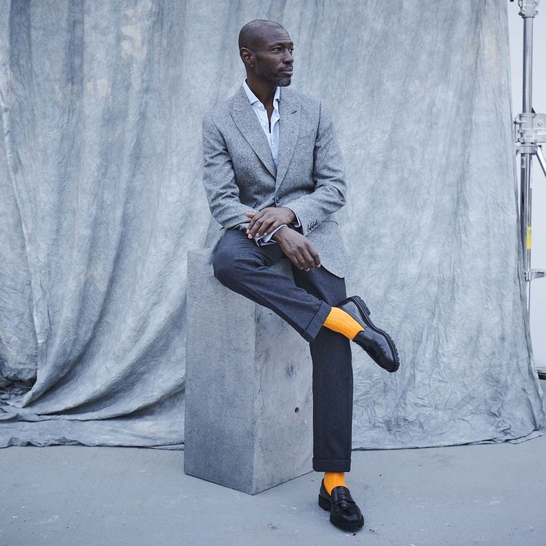 A stylish black man looks right thoughtfully, sitting on a plinth wearing charcoal grey blazer, dark grey wool trousers, black shoes and saffron socks from London Sock Company.