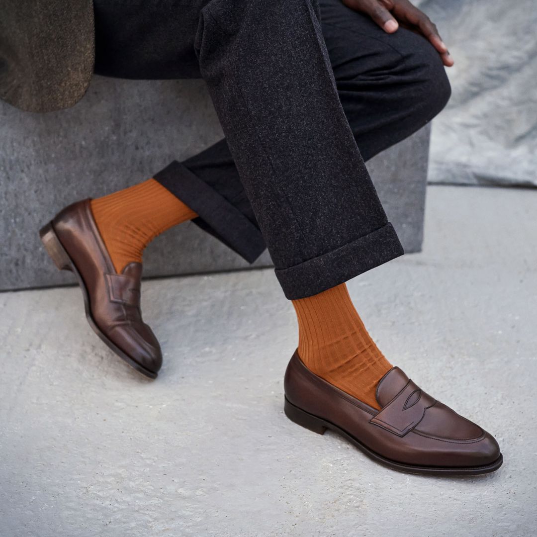 A close up of a man crossing his legs, wearing charcoal grey trousers, brown leather loafers and Rust orange socks by London Sock Company.