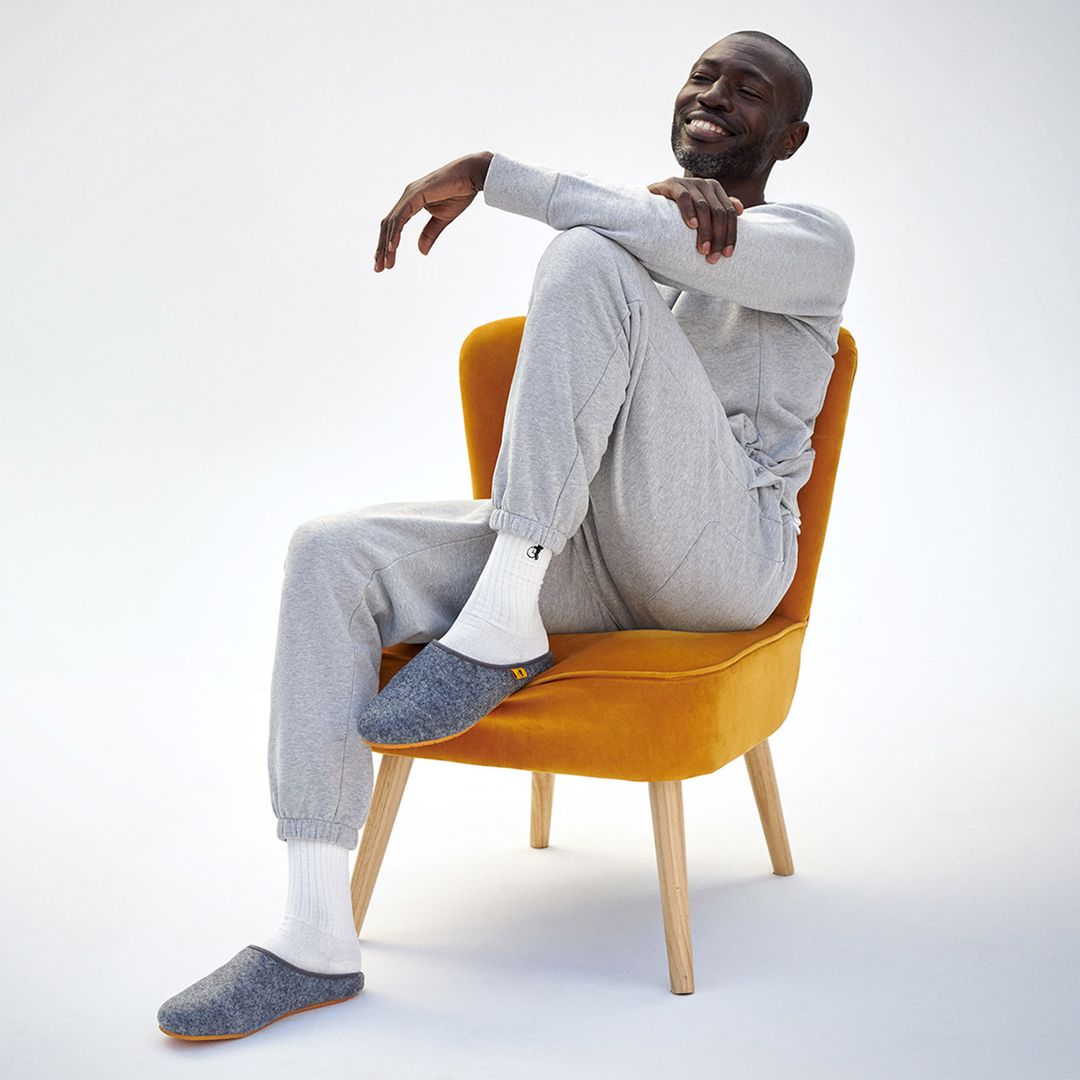 A black man in grey loungewear and white socks, smiles on a yellow chair. He is wearing London Sock Company's grey slippers.