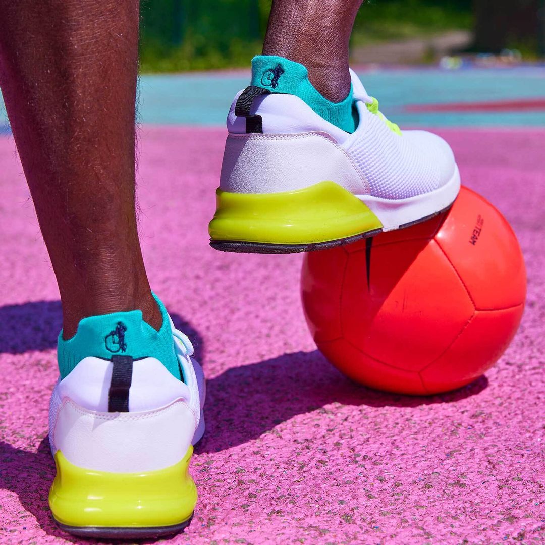 A close up of a man's feet. One foot rests on a bright pink basketball, and he is wearing white trainers and aqua ankle sports socks by London Sock Company.