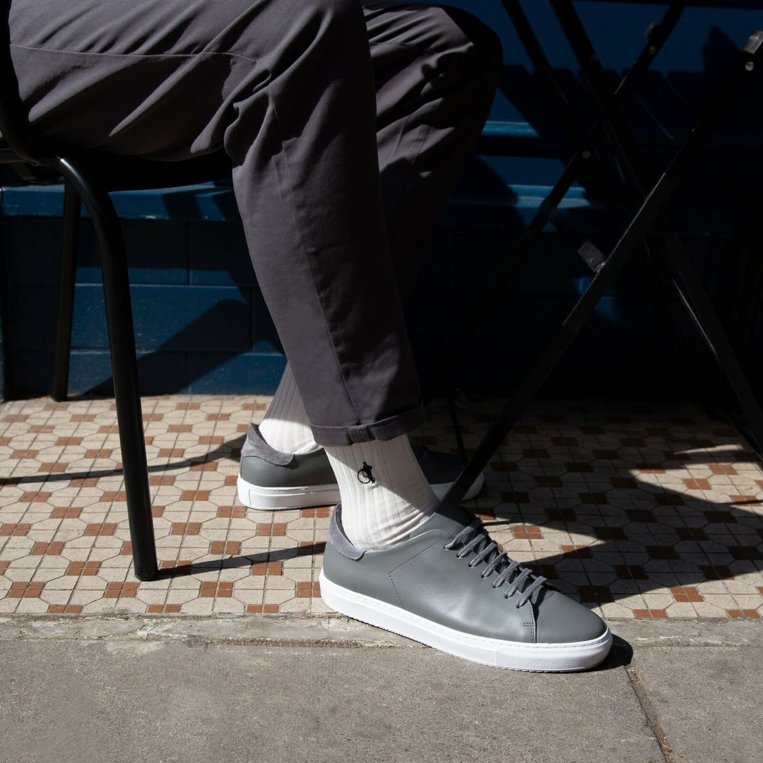 A cropped picture of a man sitting at a table in the sunshine from the knee down, his feet in plain white socks by London Sock Company and grey leather trainers.