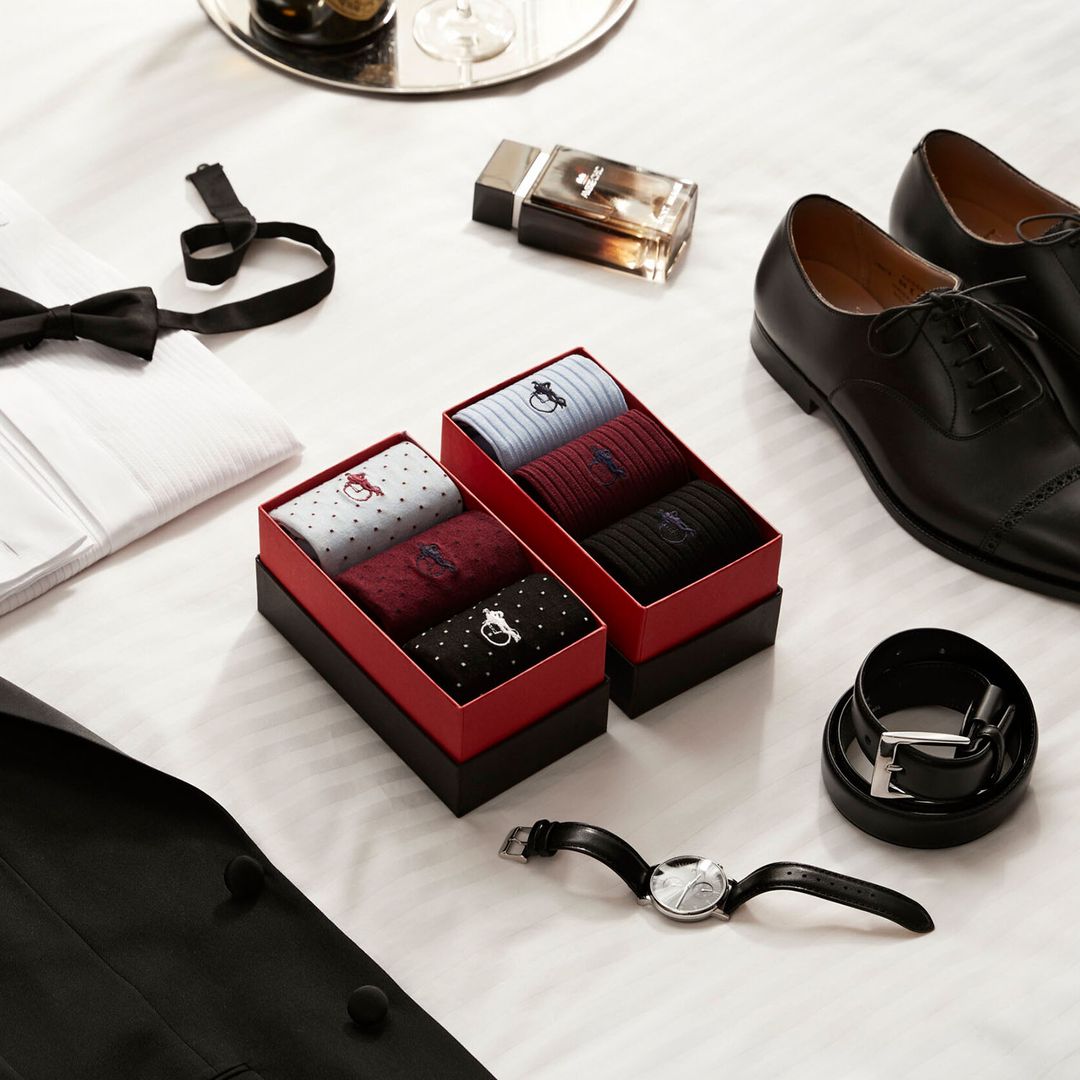 Two gift boxes of luxury socks sit on a white bedspread next to various accessories for a formal black tie look – bow tie, black oxford shoes, aftershave, and tuxedo shirt.