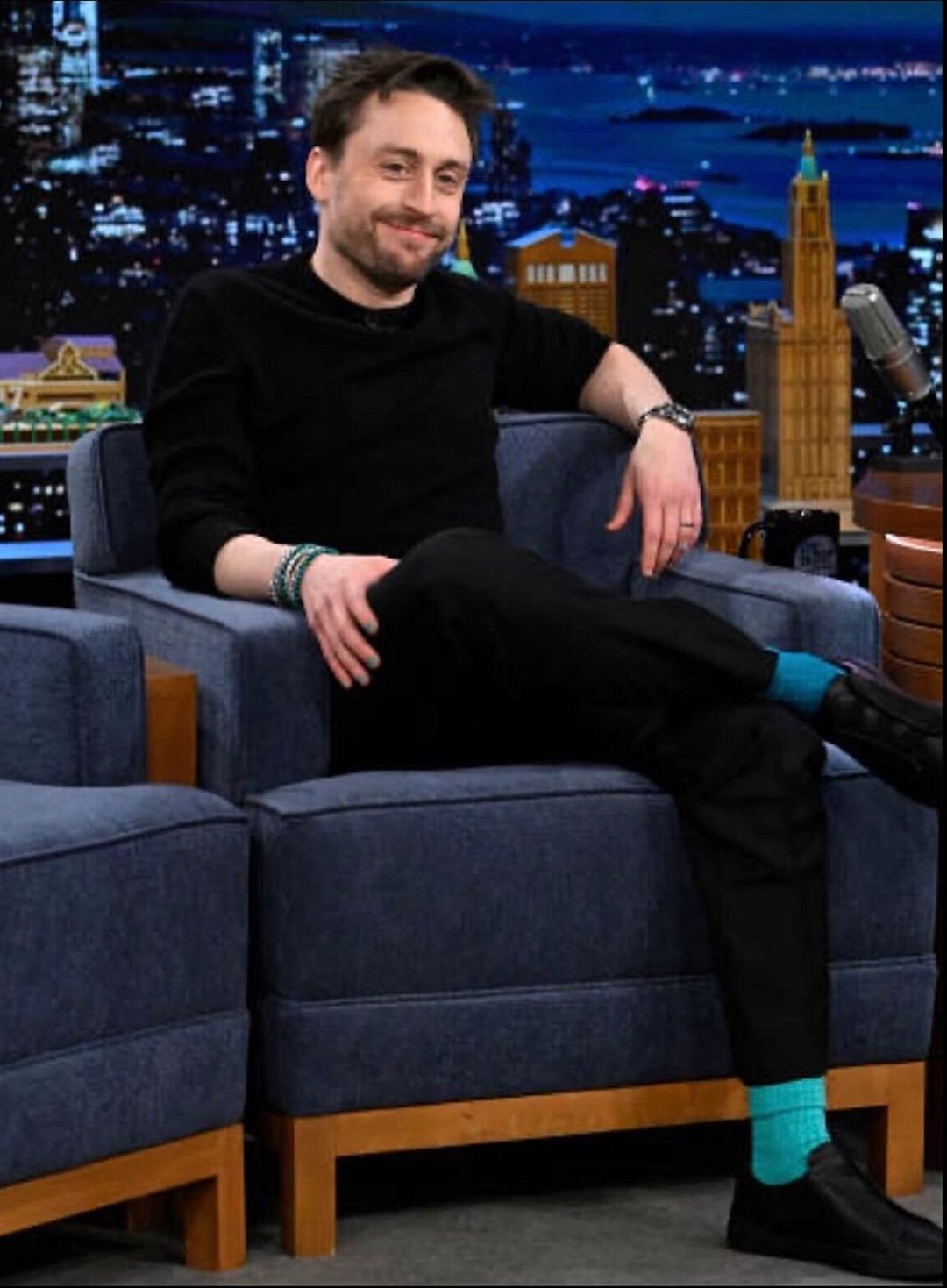 Kieran Culkin on the Tonight Show with Jimmy Fallon wearing a black tshirt and black jeans, with mismatched socks in two different shades of turquoise by London Sock Company.