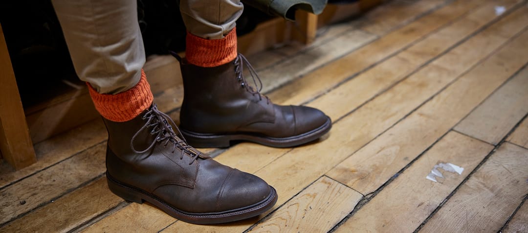 A man's feet on a wooden floor, wearing brown leather lace-up boots from Crockett & jones and spiced orange socks from London Sock Company.