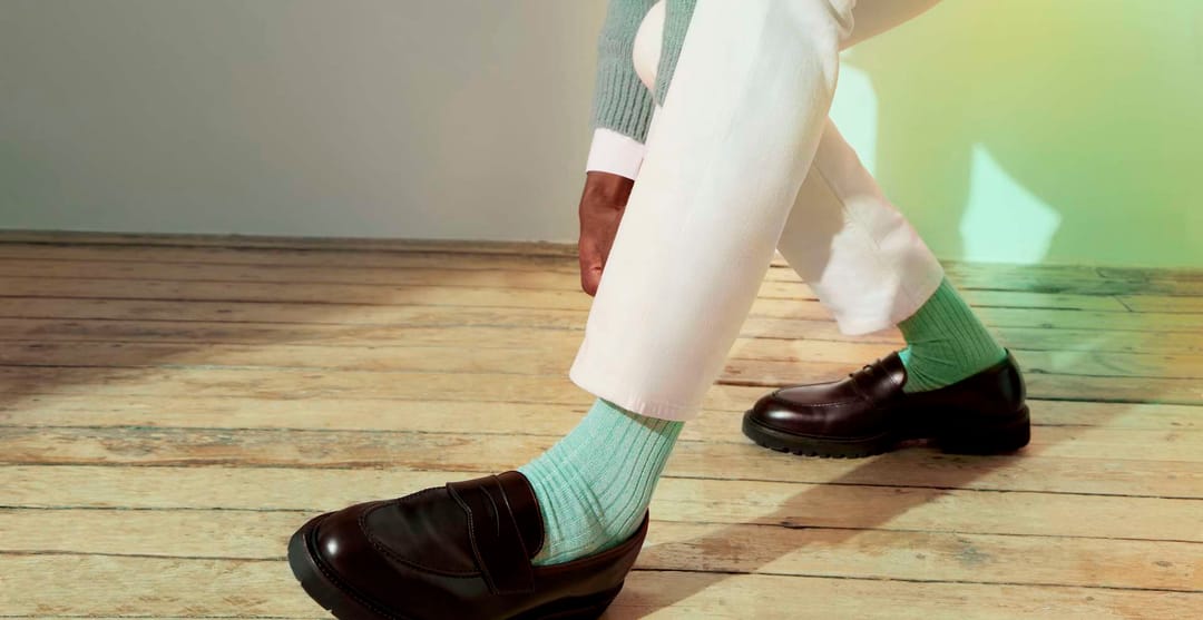 A close up of a man's feet on a hardwood floor. He is wearing black leather loafers, cream trousers, a mint green jumper and aqua green ribbed socks by the London Sock Company.