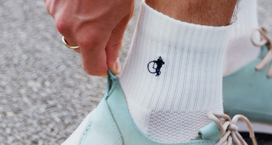 A hand pulls up the back of a blue shoe. The person wears white cropped cotton sports socks featuring London Sock Company's signature penny farthing logo embroidery.