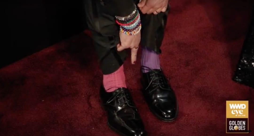 A close up of actor Kieran Culkin's feet in black dress shoes. He has colourful bracelets on his arm and he is pointing to his socks, which are mismatched – a pink sock from London Sock Company on the right foot and a purple sock from London Sock Company on the left foot. 