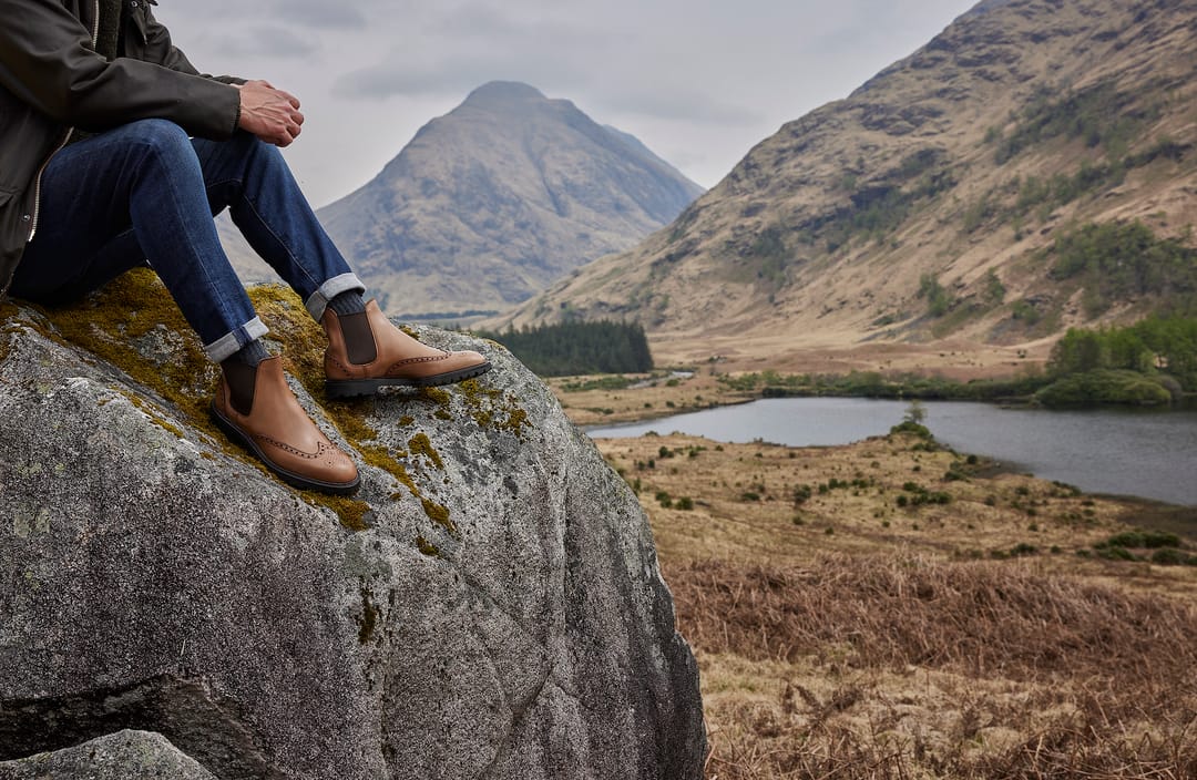 A man sits on a rock with a view of a Highland glen behind him, wearing jeans, Chelsea boots and grey boot socks by London Sock Company.