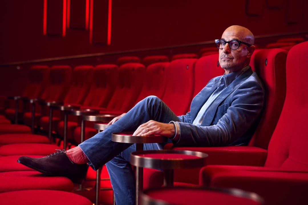 Actor Stanley Tucci sits in classic red theatre chair wearing coral pink socks by London Sock Company.