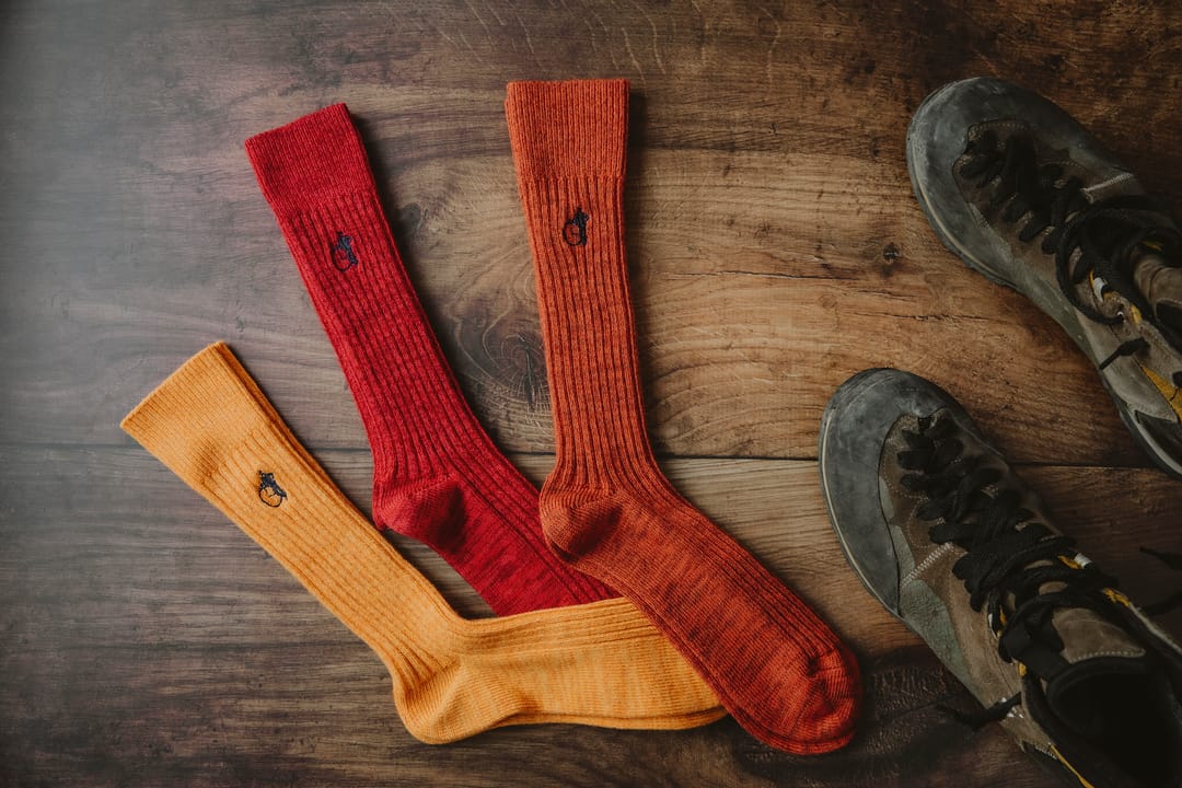 A flatlay image of a pair of yellow, a pair of red and a pair of orange boot socks by London Sock Company on a wooden floor next to some hiking boots. 