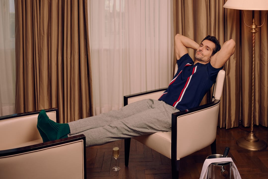 A man sits in a chair looking at the camera, with his arms behind his head and his feet up on another chair. He is wearing a blue knitted polo, beige smart trousers, and green merino wool socks from London Sock Company.