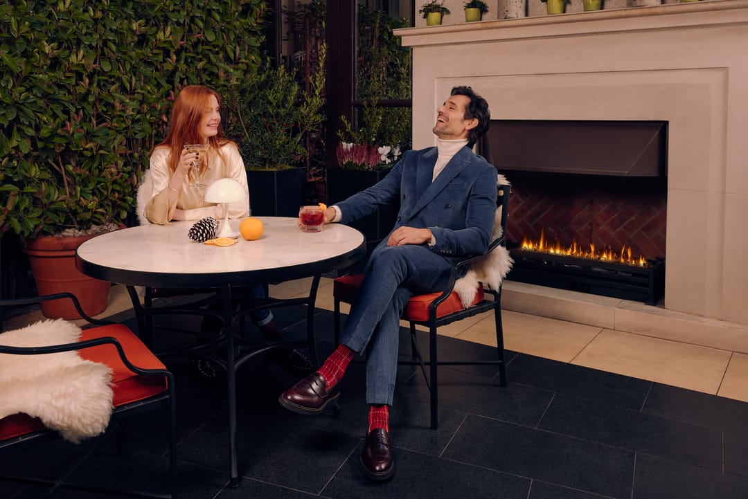 A man and woman sitting at a table in front of a fireplace, the man is wearing London Sock Company's Ottaway Style Socks in a deep red colour.