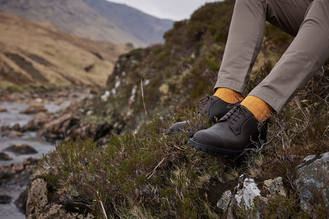 A man sits in a Scottish glen, surrounded by heather with a stream in the background, wearing yellow boot socks by London Sock Company and brown leather shoes by Crockett & Jones.
