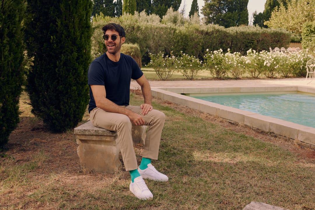 A man with dark hair sits on a stone bench among cyprus trees wearing a navy tshirt, tan chinos and white trainers, with aqua coloured socks by London Sock Company.
