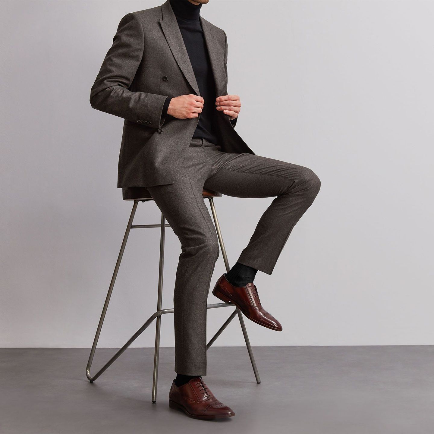 Man sitting in dark grey suit and black socks a matching top and brown shoes