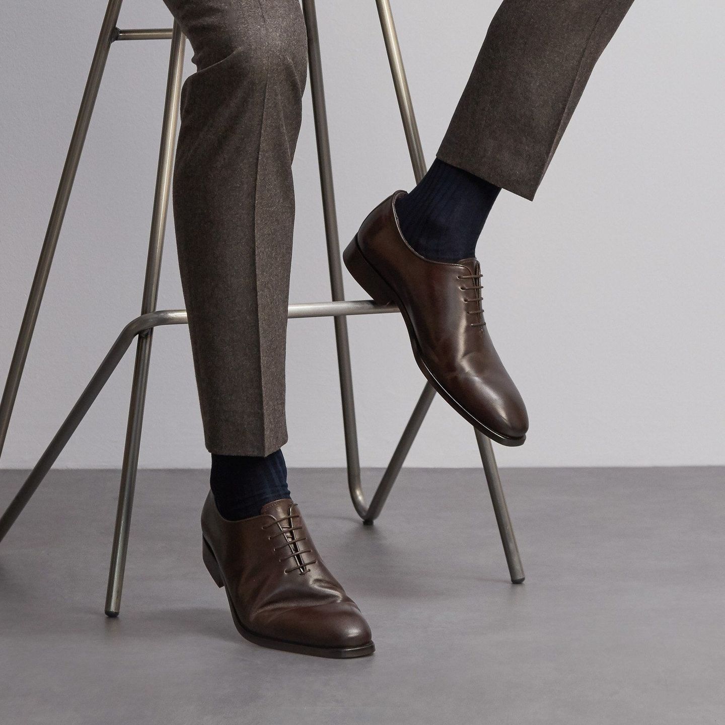 Close up of a man's legs sitting in a grey suit, navy sartorial socks and brown shoes