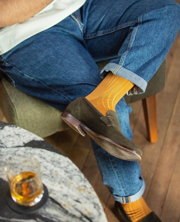 A man in jeans crossed-legged wearing mustard yellow socks with a dram of whisky by his side.